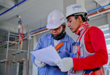 two men in hard hats look at white paper together